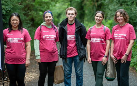 Amy, Georgia, Cree, Bryony and Lucy stood in a line on the woodland pathway smiling at the camera. Everyone is wearing a pink YMCA DownsLink Challenge t-shirt