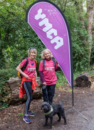 Nikki, Julia, and her Dog Bruce smiling next to a pink YMCA flag on the woodland pathway