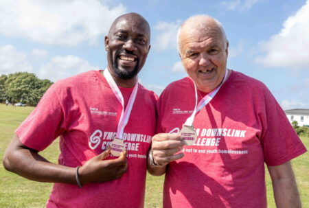 Dennis and Geordie smiling at the camera, holding up their finisher medals. They are both also wearing a pink YMCA DownsLink Challenge t-shirt