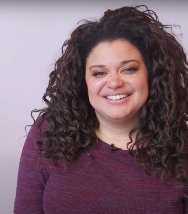Michelle Buteau smiling at the camera wearing a purple tshirt. Black history month