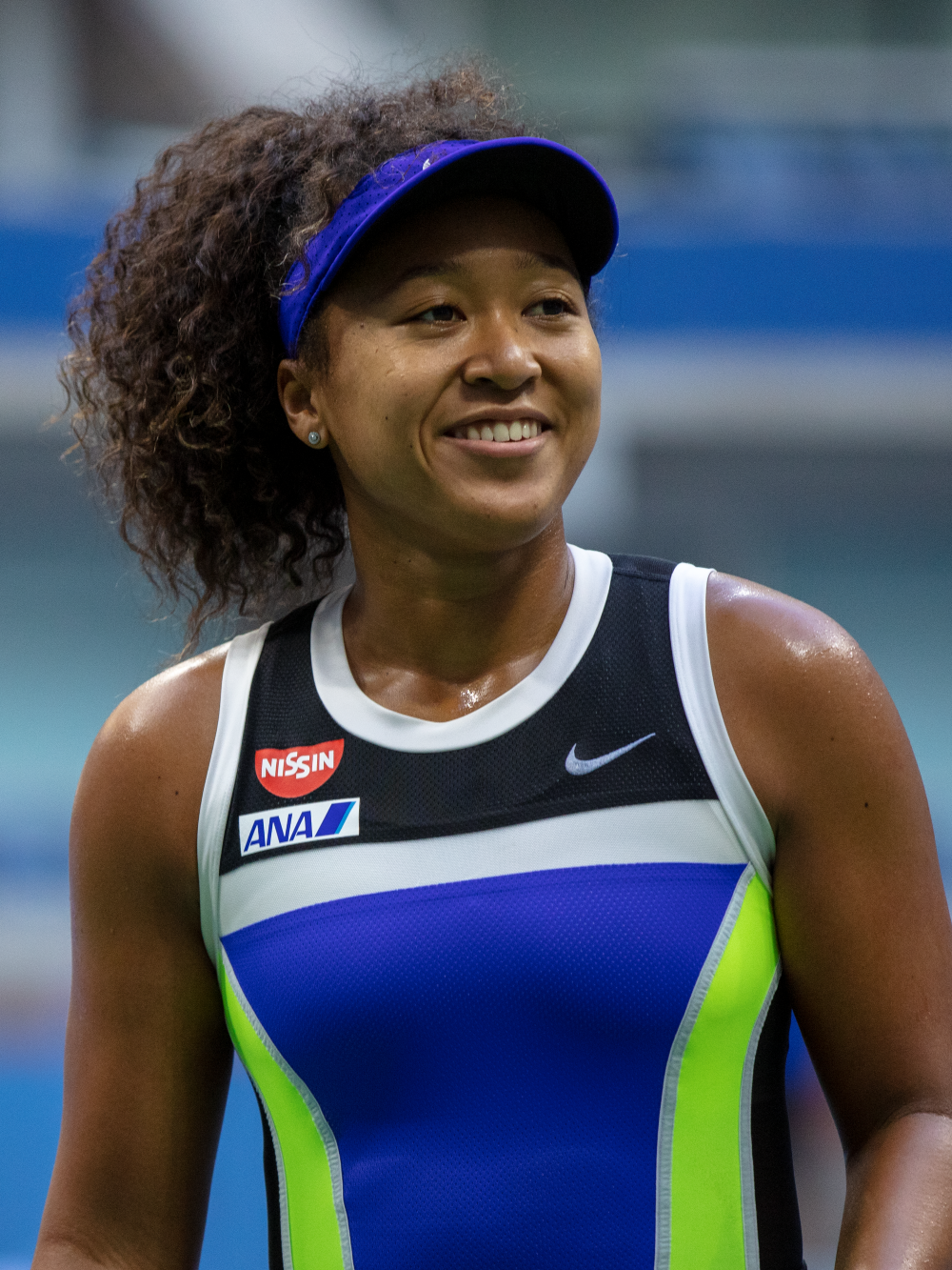 Naomi Osaka playing tennis wearing a blue cap and black and blue sport vest. She is smiling to the right. Black history month